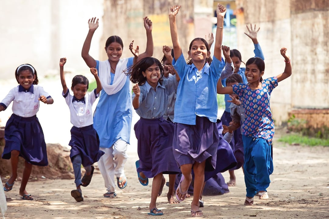 Group of young Indian girls running with their hands in the air, smiling in school uniforms.